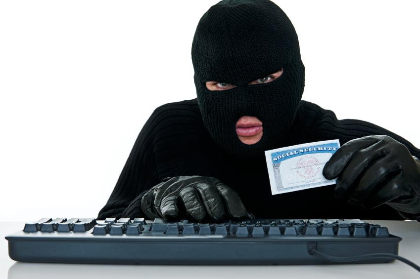 Masked identity thief wearing gloves holding social security card with right hand on computer keyboard.
